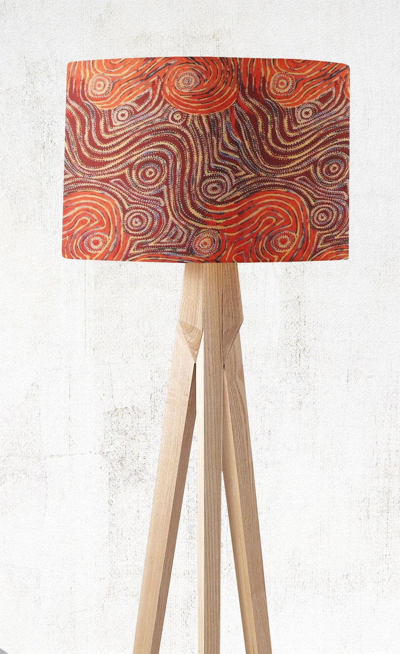 The Indigenous Aboriginal Warlu Mina Mina Dreaming Lampshade by artist Pauline Napangardi Gallagher features an abstract design with orange, red, and white swirls and dots on a light textured background atop a wooden tripod floor lamp.