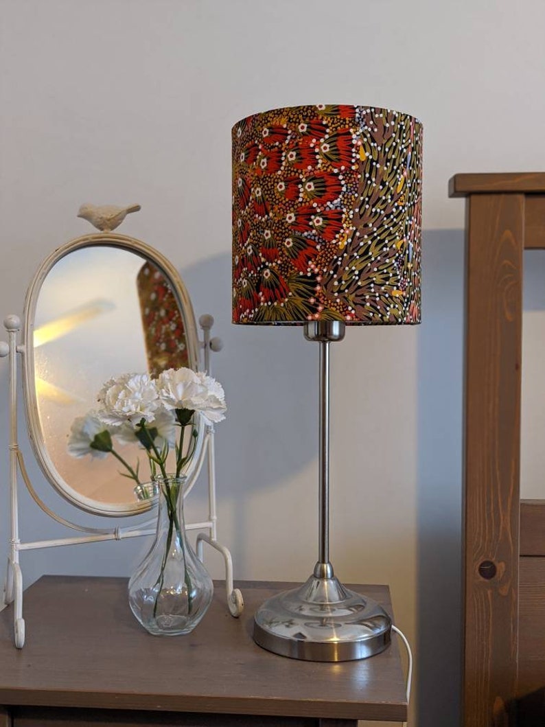 A bedside table with the Bush Banana Lampshade by artist Donna Abbot, featuring a colorful Indigenous-inspired design, plus a silver lamp base, white flowers in a glass vase, and an oval mirror with a bird accent. A wooden bed frame is partly visible.
