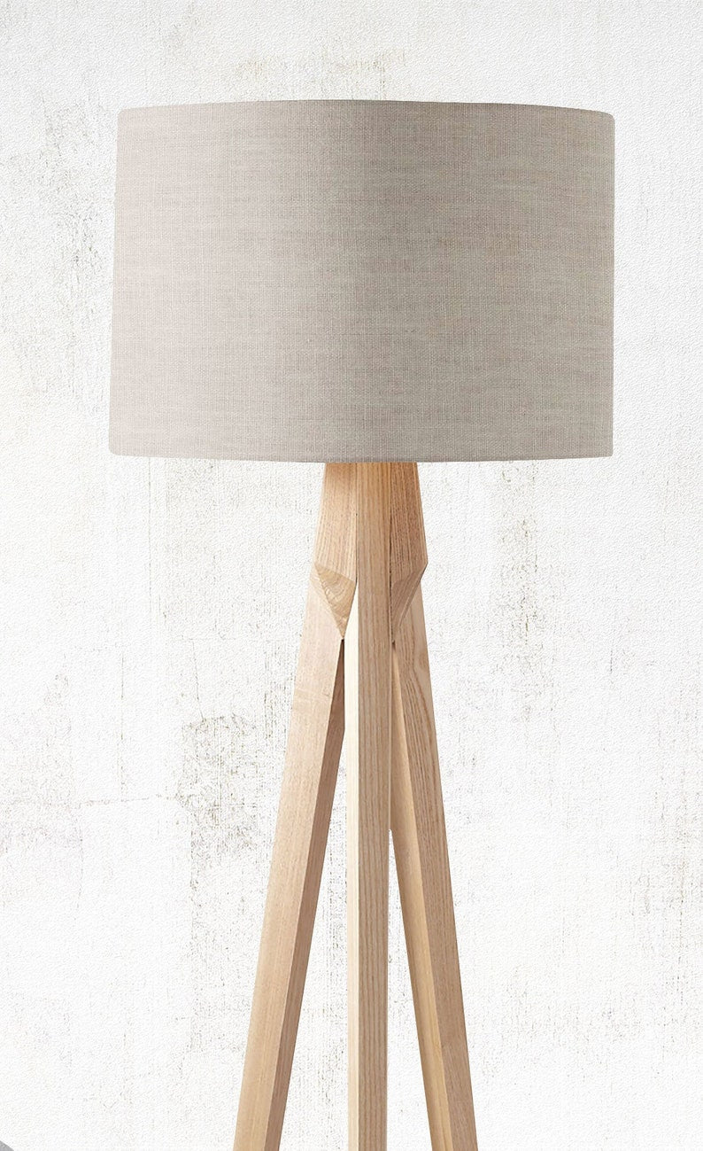 The Emotive Collection’s Stone Premium Linen Lampshade features a large round linen shade and a modern light wood tripod base, beautifully set against a white textured wall.
