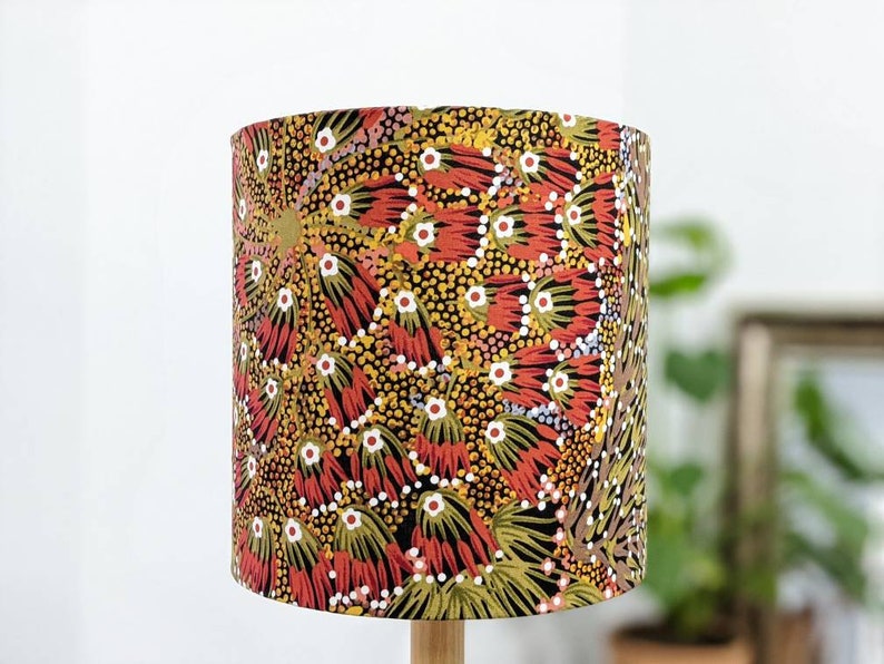 The Bush Banana Lampshade by artist Donna Abbot features vibrant red, yellow, and green brush-like patterns with white dots inspired by Indigenous art, shown indoors with a blurred plant and picture frame in the background.