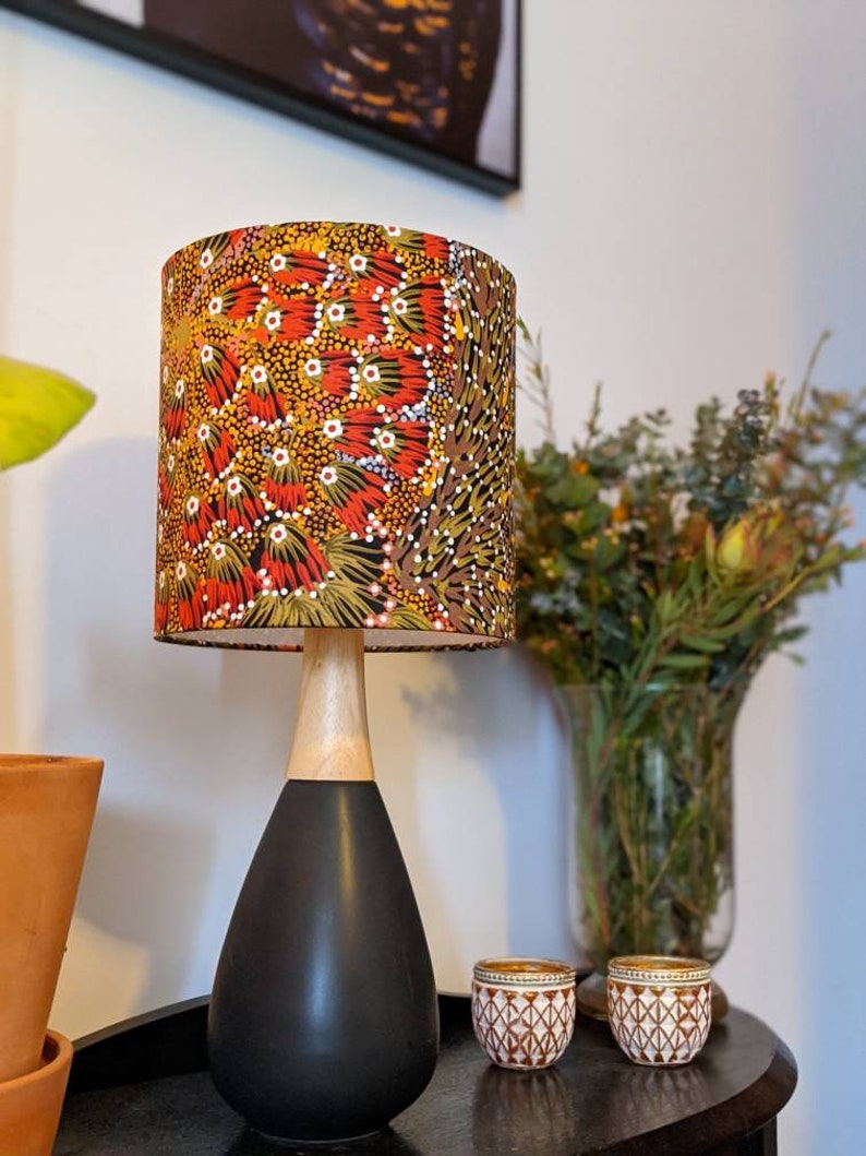 A sleek black and wood table lamp with the Bush Banana Lampshade by artist Donna Abbot sits on a dark surface beside two decorative jars, with a vase of greenery and a terracotta pot in the background.