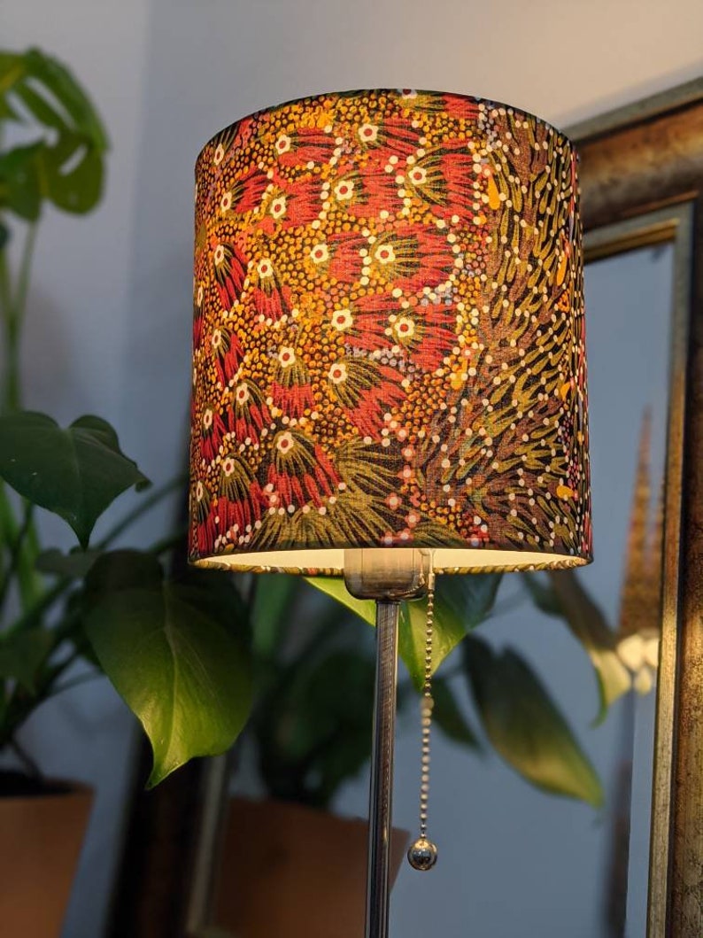 A standing lamp with the Bush Banana Lampshade by artist Donna Abbot, showcasing a vibrant Aboriginal design in red, yellow, and brown, stands beside green plants and a framed mirror for a cozy, stylish touch.