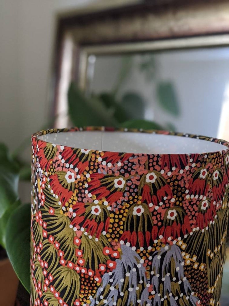 A close-up of the Bush Banana Lampshade by Artist Donna Abbot, showing its vibrant Aboriginal-inspired floral pattern in red, green, black, and white, set before a blurred mirror and leafy plant.
