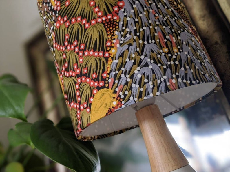 A close-up of a table lamp featuring the Bush Banana Lampshade by artist Donna Abbot, with Indigenous art in red, yellow, white, and gray. The lamp has a wooden base and a blurred green plant in the background.