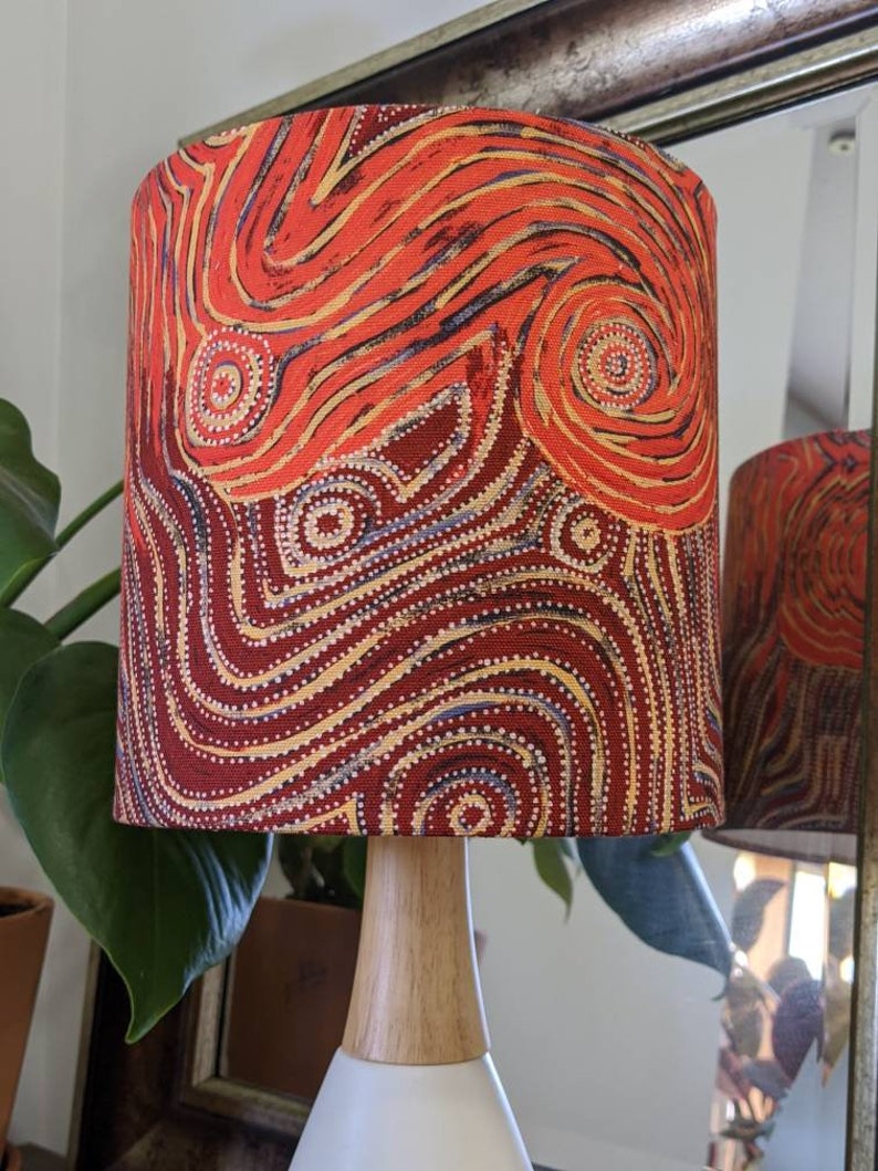 Next to a green plant and a mirror stands the Indigenous Aboriginal Warlu Mina Mina Dreaming Lampshade by Pauline Napangardi Gallagher, featuring vibrant red, orange, and yellow swirling and dotted designs on a wooden base.