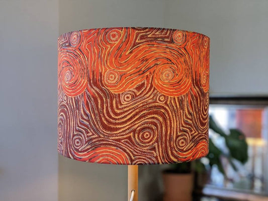 The Indigenous Aboriginal Warlu Mina Mina Dreaming Lampshade by artist Pauline Napangardi Gallagher features swirling red, orange, and brown lines on a handmade shade with a wooden base; a blurred potted plant is seen in the background.