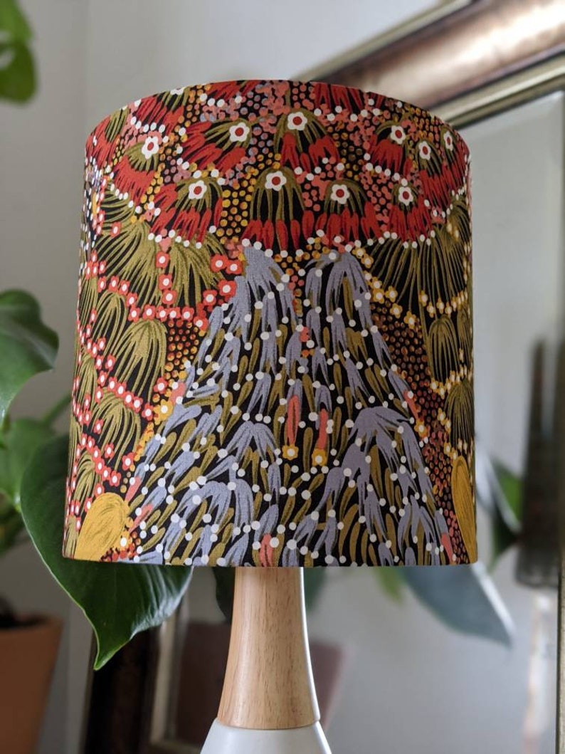 The Bush Banana Lampshade by artist Donna Abbot features a wooden base and a colorful, abstract floral shade inspired by Aboriginal designs in red, gold, green, and purple. A large leaf and part of a mirror appear in the background.