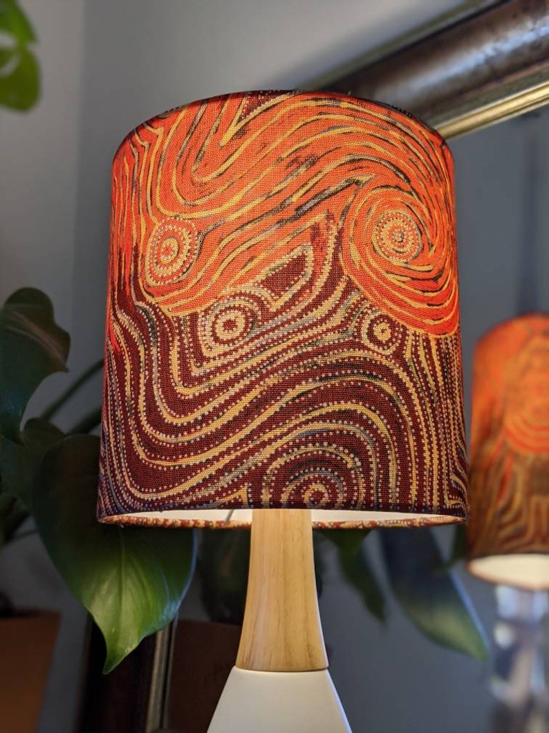 The Indigenous Aboriginal Warlu Mina Mina Dreaming Lampshade by artist Pauline Napangardi Gallagher, with a wooden base and handmade shade in vibrant red, orange, and yellow patterns, sits beside a mirror and lush green plant leaves.