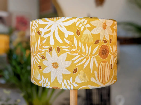 The Native Flora Mustard Lampshade by Emotive Collection features Jocelyn Prousts bold white and brown floral design on yellow, shown indoors with blurred plants and furniture in the background.