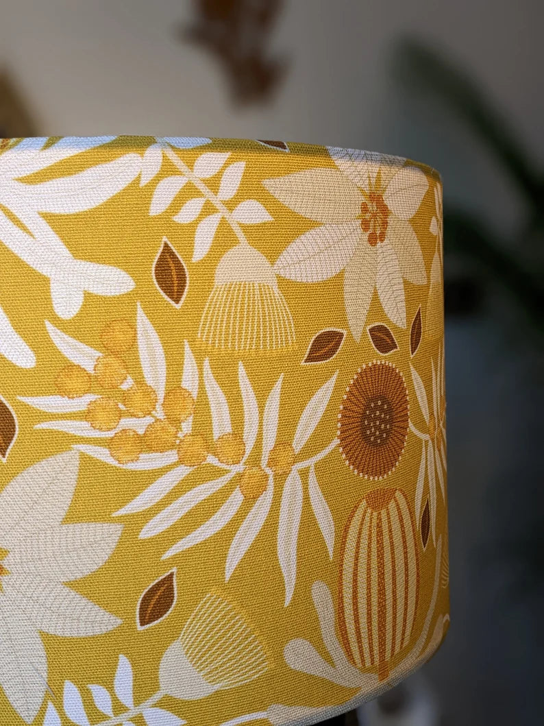 The Emotive Collections Native Flora Mustard Lampshade features a handmade yellow design with Jocelyn Proust’s white, brown, and orange floral pattern—leaves, flowers, and seeds vivid on a soft blurred backdrop.