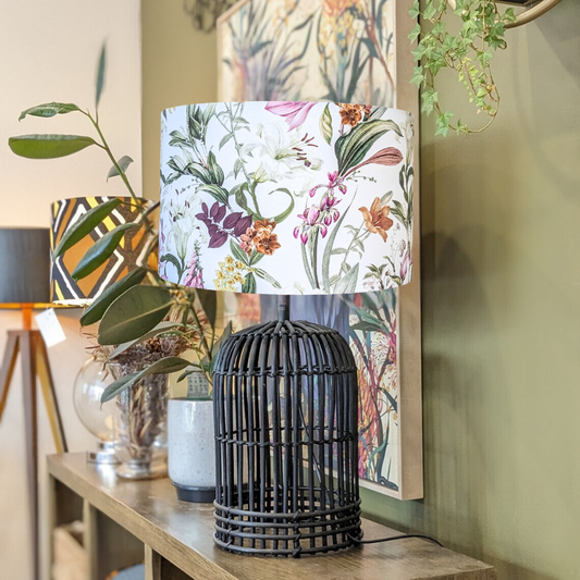 The Emotive Collection Summer Floral Lampshade sits on a black wicker table lamp atop a wooden console, surrounded by green plants, decor accents, and botanical wall art in the background.