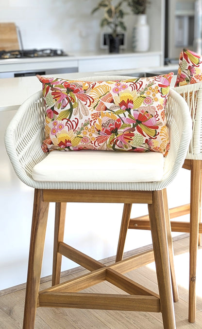 A wooden bar stool with a white woven backrest and seat cushion features an Emotive Collection Wild Protea Cushion Cover. The cotton canvas cover adds color to the bright, modern kitchen. Another matching stool is partially visible nearby.