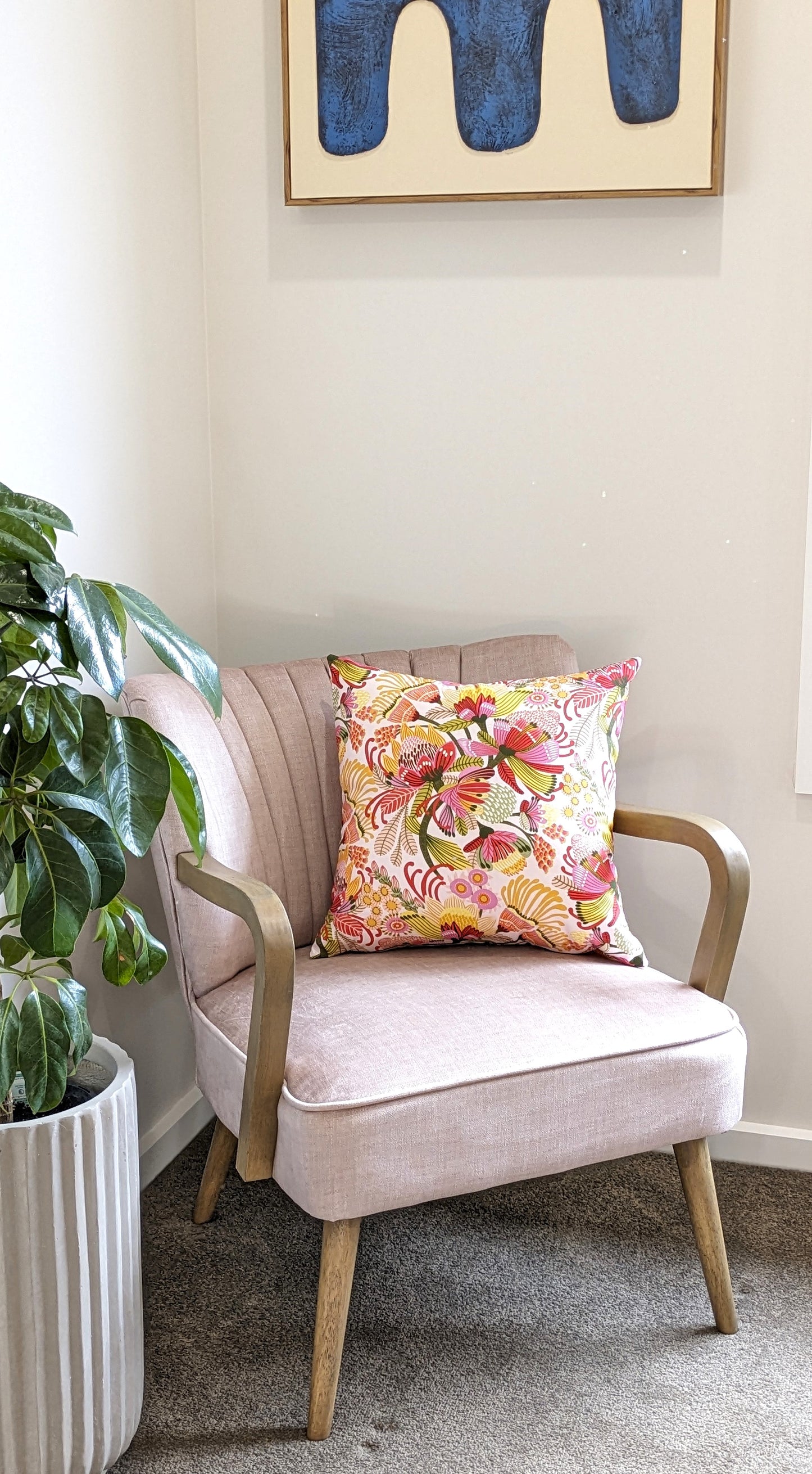 A light pink armchair with wooden legs and arms sits in a corner, topped with the Emotive Collections Wild Protea Cushion Cover. A green potted plant is on the left, and abstract blue artwork by Kirsten Katz hangs above.