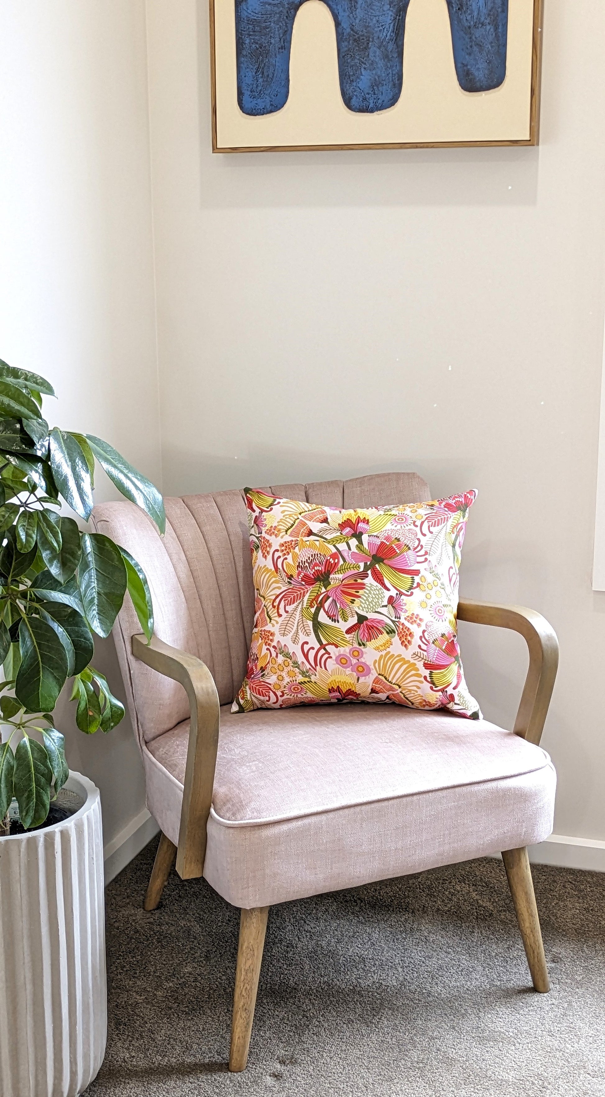 A light pink armchair with wooden legs and arms sits in a corner, topped with the Emotive Collections Wild Protea Cushion Cover. A green potted plant is on the left, and abstract blue artwork by Kirsten Katz hangs above.