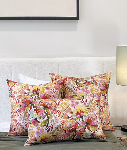 Three vibrant Wild Protea Cushion Covers by Emotive Collection, crafted from cotton canvas, are displayed on a white upholstered bed with a white pillow behind them; a lamp and wall art can also be seen in the background.