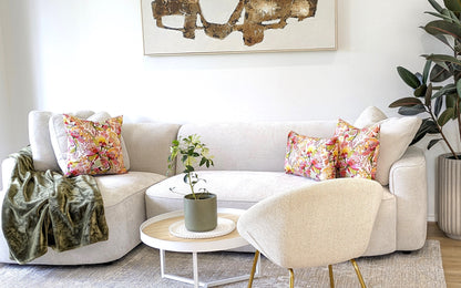 A modern living room with a white curved sofa in cotton canvas, Wild Protea Cushion Cover by Emotive Collection, a green throw, round coffee table with potted plant, abstract Kirsten Katz wall art, and a large leafy plant in the corner.