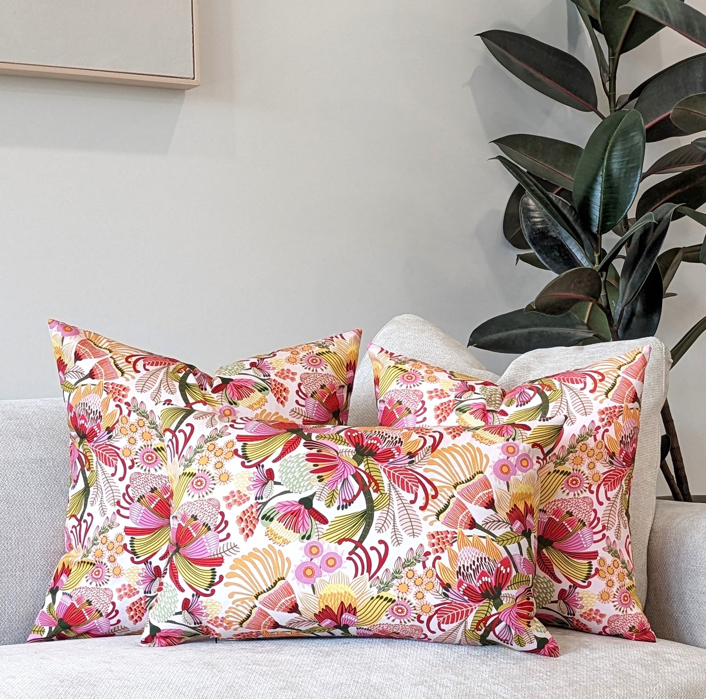Three Emotive Collection Wild Protea Cushion Covers, featuring vibrant Kirsten Katz floral designs on cotton canvas, brighten a light sofa. A leafy plant and framed picture complete the neutral setting.