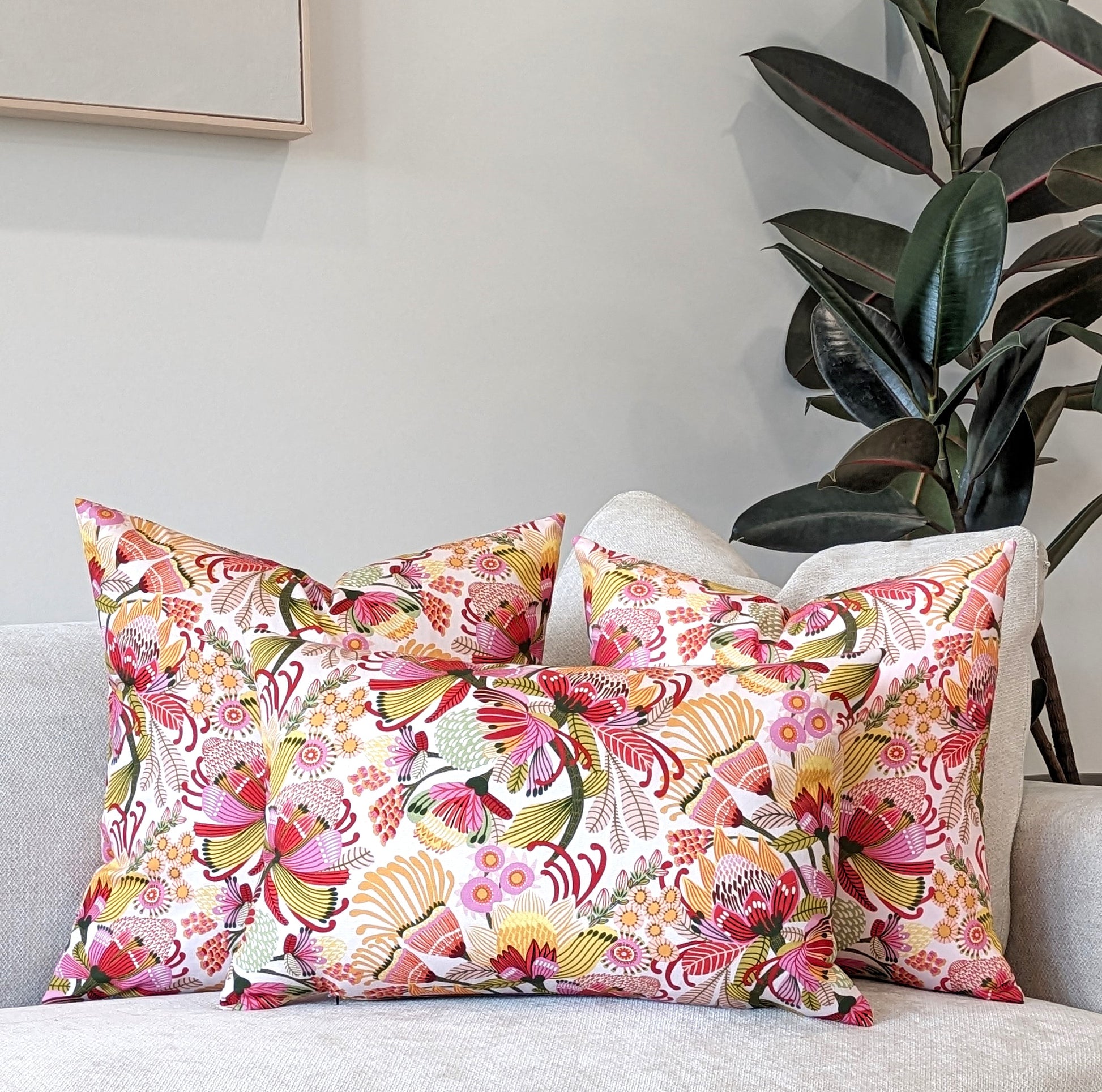 Three Emotive Collection Wild Protea Cushion Covers, featuring vibrant Kirsten Katz floral designs on cotton canvas, brighten a light sofa. A leafy plant and framed picture complete the neutral setting.