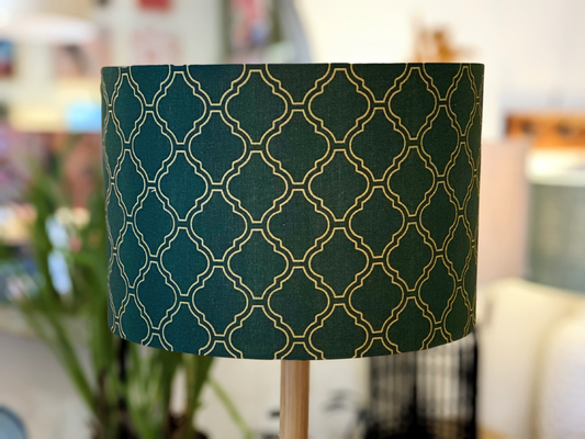 A close-up of the Art Deco Lampshade (35cm x 24cm) by Art Deco, featuring dark green fabric with a gold geometric pattern, set indoors with softly blurred plants and furniture in the background.