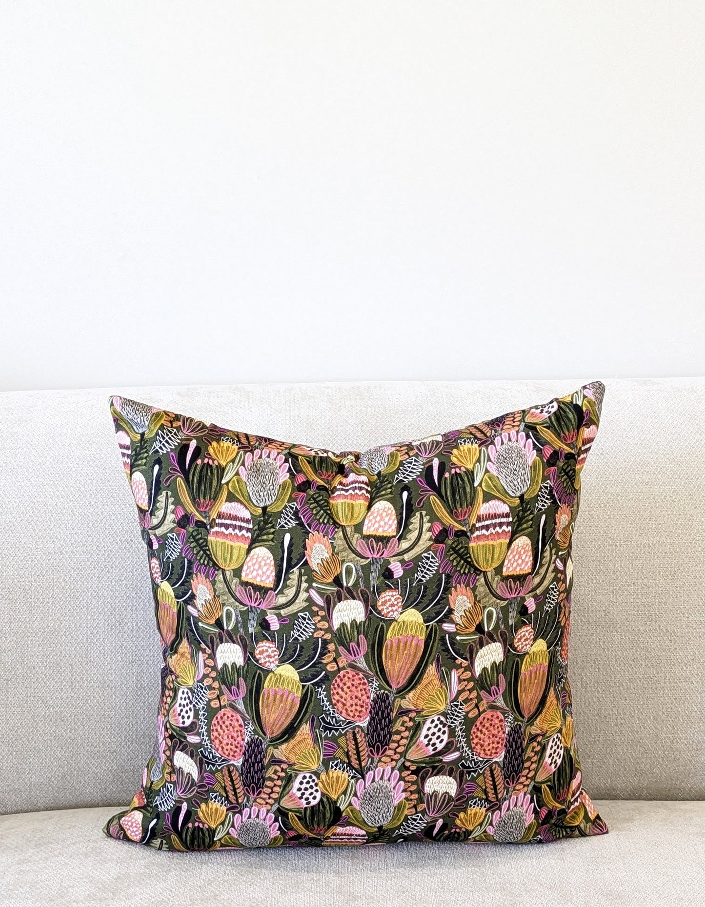 The Emotive Collection’s Bush Banksia Cushion Cover, featuring Kirsten Katzs vibrant design, is displayed on a light beige sofa against a plain white wall.