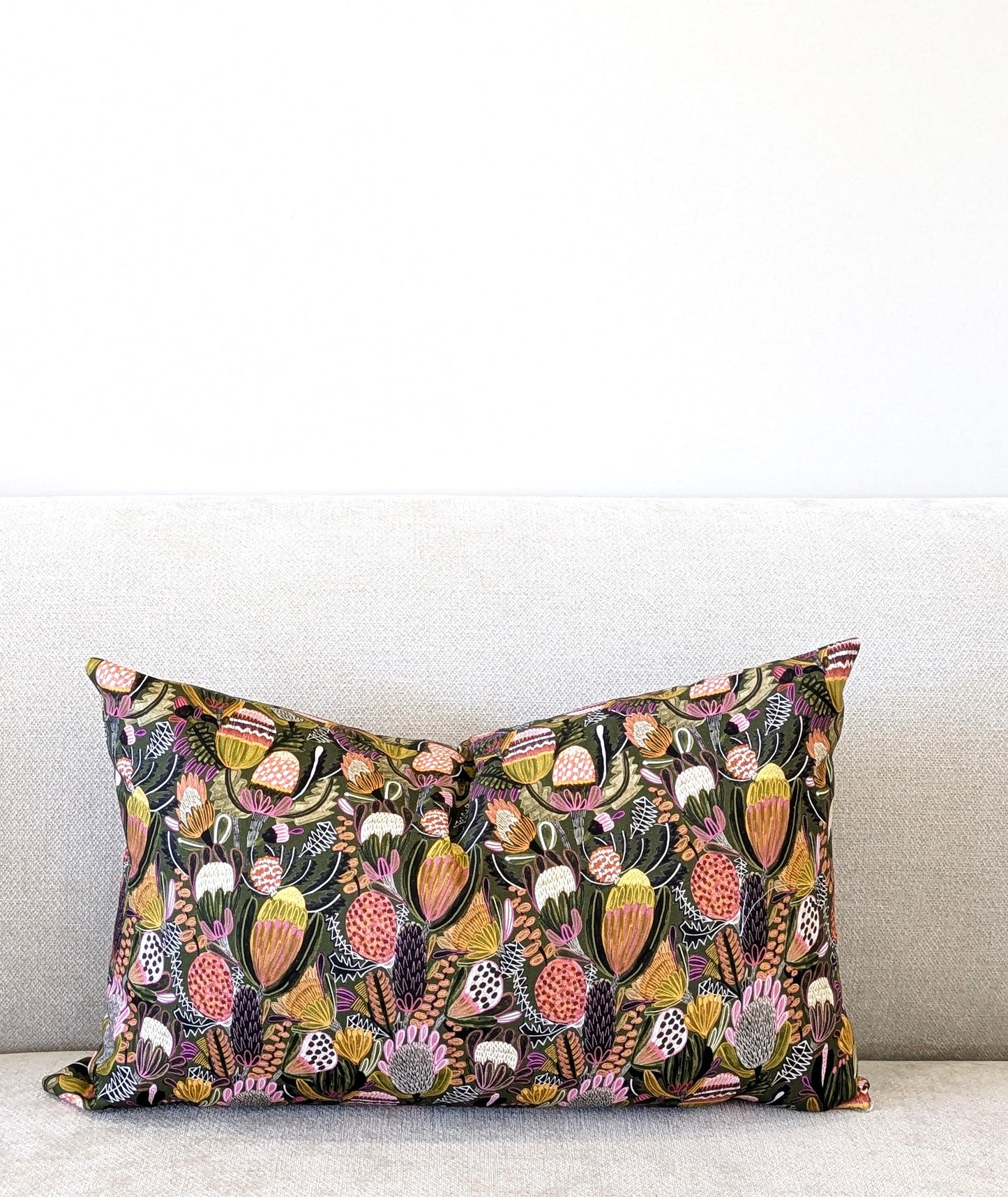 The Emotive Collection Bush Banksia Cushion Cover, crafted from cotton canvas and featuring a colorful floral pattern, is styled on a light beige upholstered sofa set against a plain white wall.