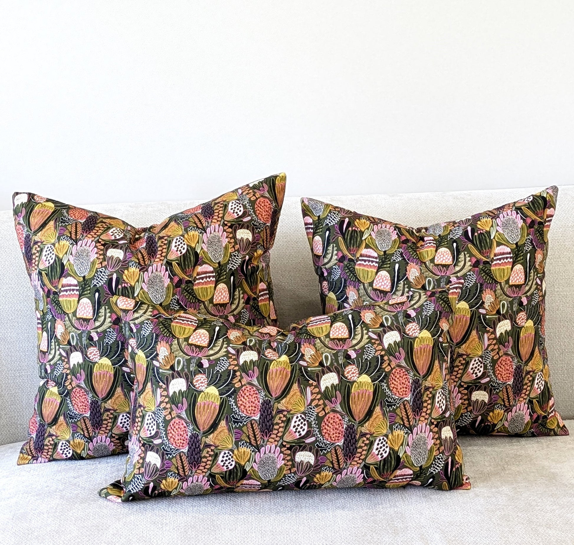 Three Bush Banksia Cushion Covers from the Emotive Collection, featuring vibrant floral and botanical patterns in pink, orange, green, and yellow, are displayed on a light sofa set against a plain white wall.