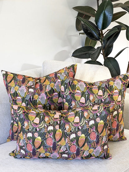 Three Emotive Collection Bush Banksia Cushion Covers add charm to a light sofa with their colorful botanical design, while a dark green rubber plant stands against the pale wall in the background.