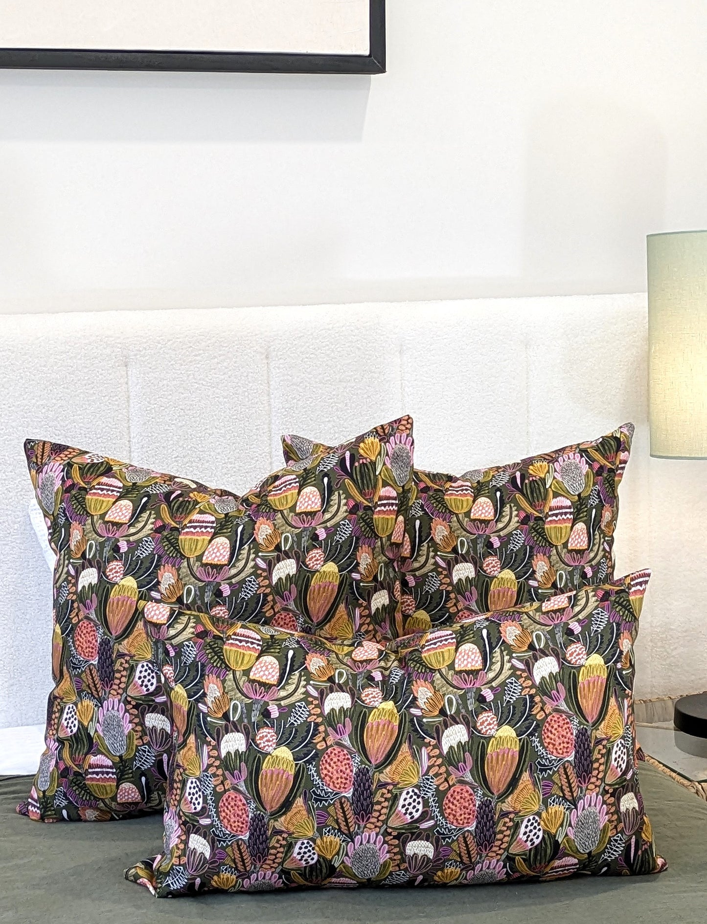 Four Emotive Collection Bush Banksia Cushion Covers in vibrant floral prints are arranged on a bed with a white headboard. A softly lit background reveals a small lamp and part of a picture frame.