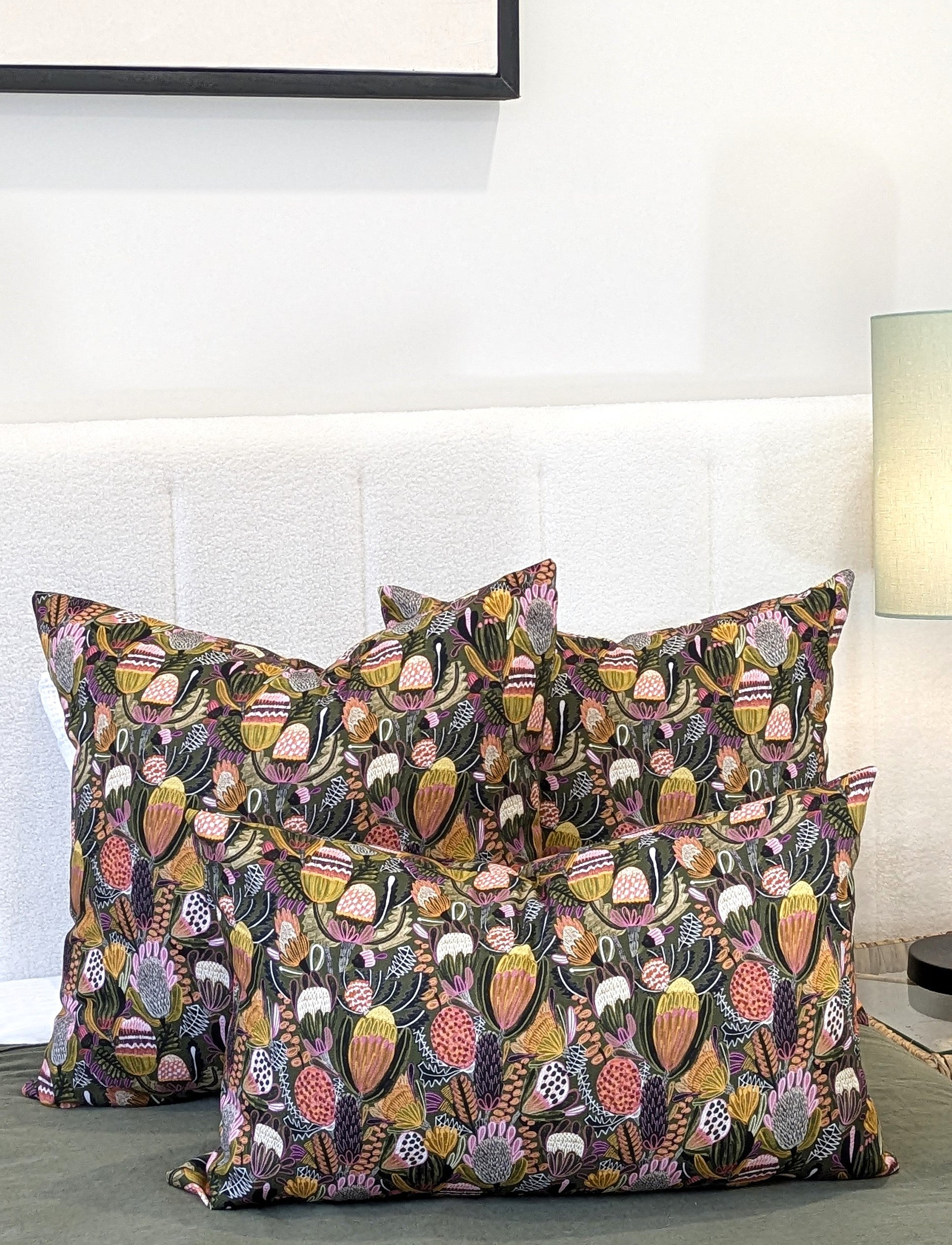 Four Emotive Collection Bush Banksia Cushion Covers in vibrant floral prints are arranged on a bed with a white headboard. A softly lit background reveals a small lamp and part of a picture frame.