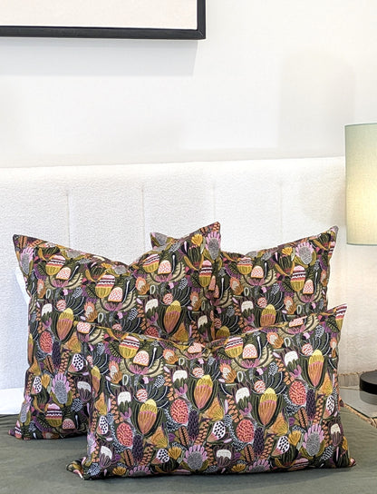 Four Emotive Collection Bush Banksia Cushion Covers in vibrant floral prints are arranged on a bed with a white headboard. A softly lit background reveals a small lamp and part of a picture frame.