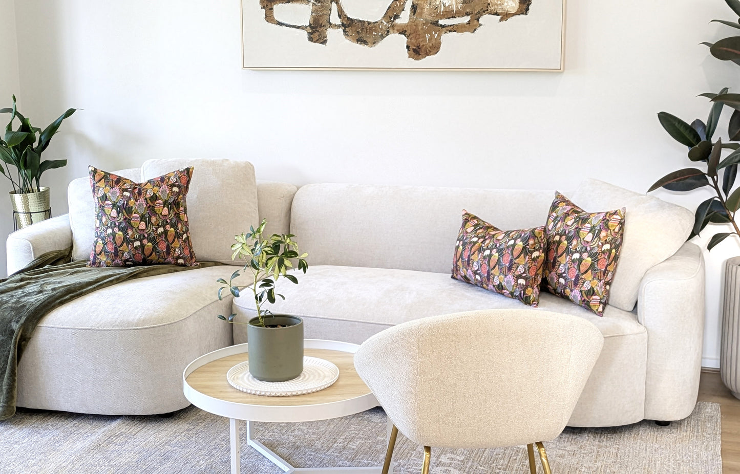 A modern living room with a light beige sofa featuring Emotive Collection’s Bush Banksia Cushion Cover, colorful patterned pillows, a round white coffee table with a potted plant, cream accent chair, abstract wall art, and green plants.