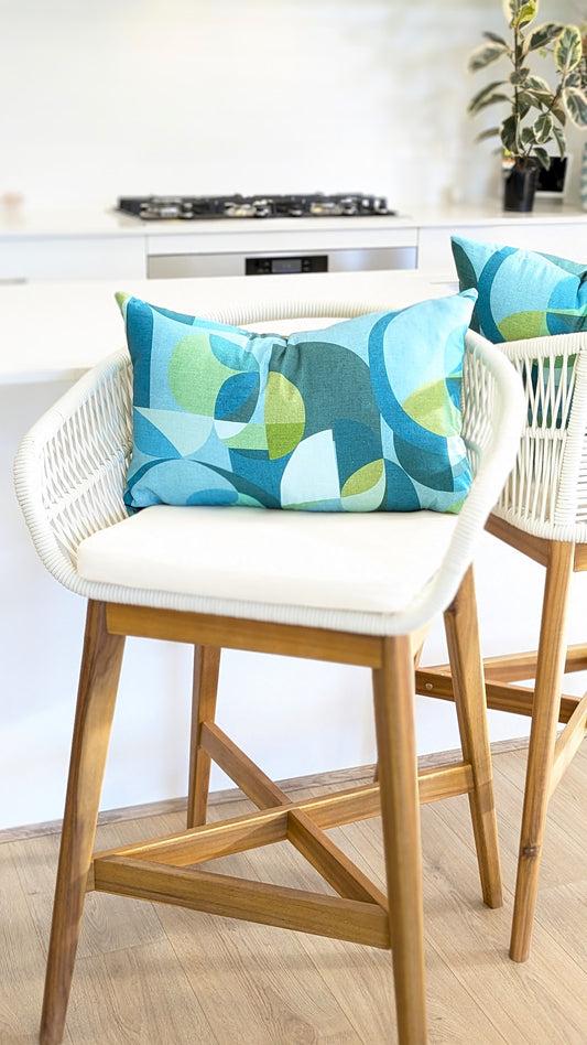 A white barstool with wooden legs sits by a kitchen counter, topped with a handmade linen cushion and an Emotive Collection Linen Mid Century Blue Cushion Cover. Another similar stool is partly visible nearby.