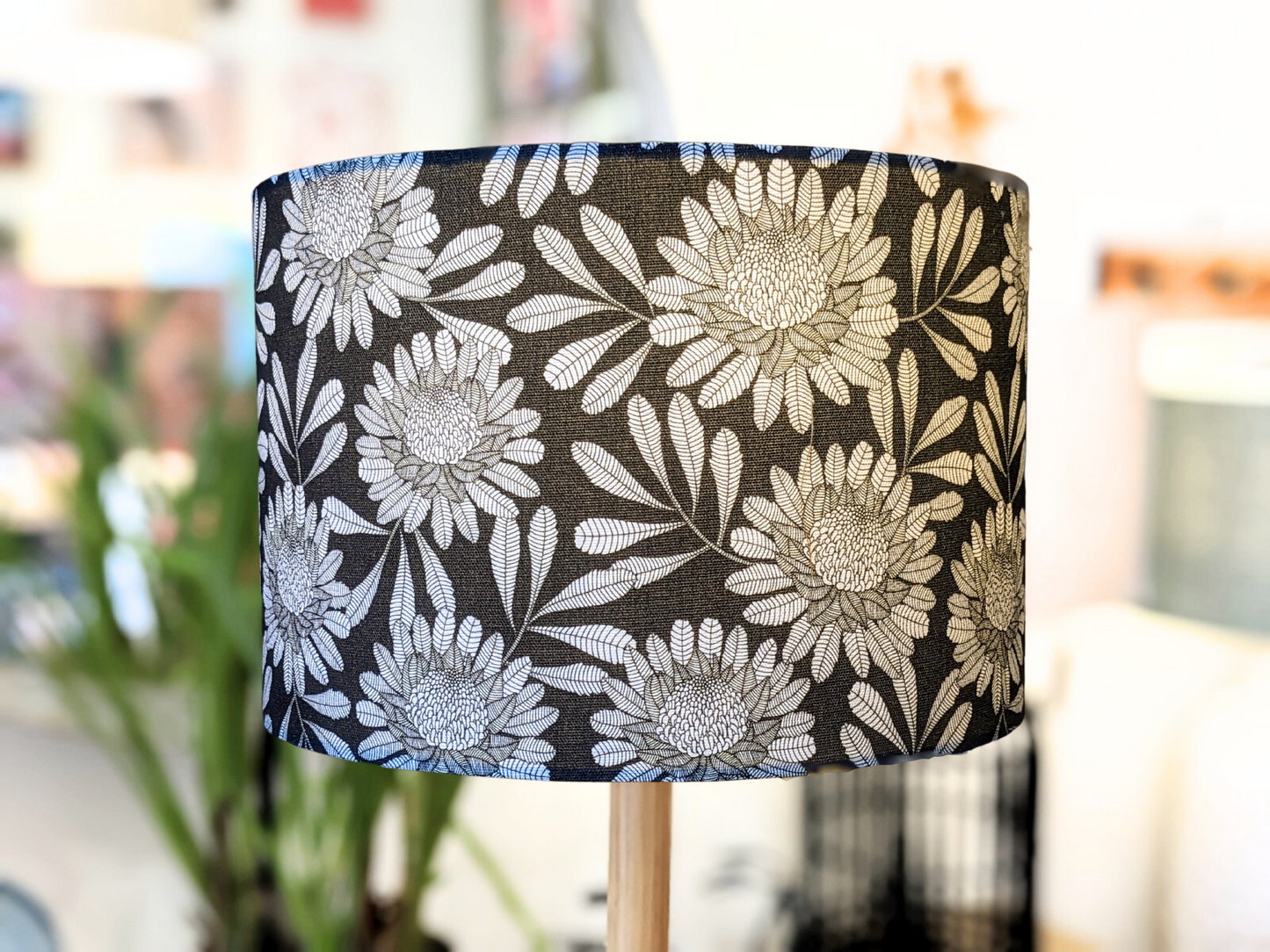 A close-up of the Emotive Collection’s Australian Waratah Lampshade, handmade with a bold white floral pattern on black, large flowers and leaves in Jocelyn Prousts style. The lamp is indoors, background softened with plants and furniture.