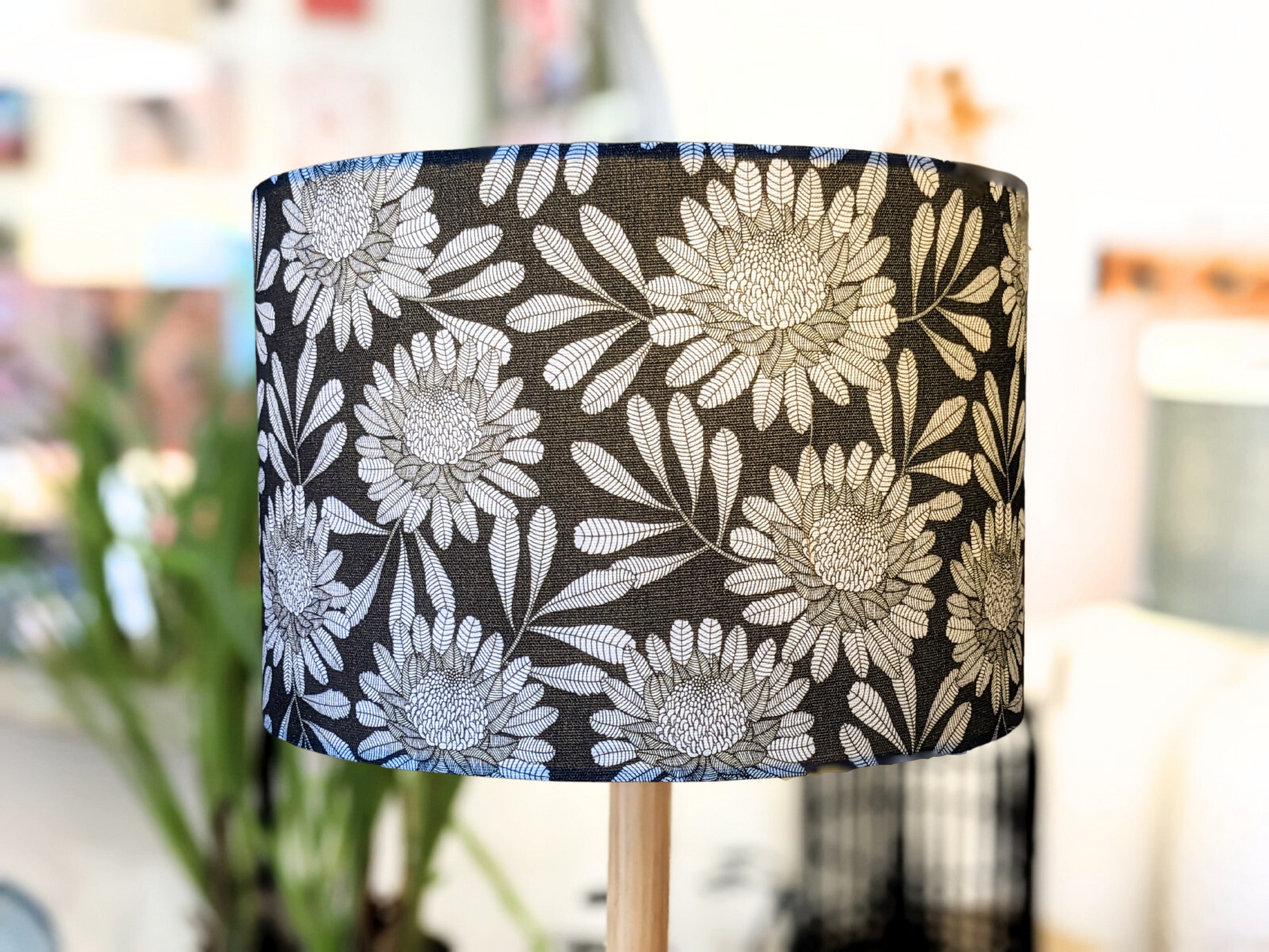 A close-up of the Emotive Collection’s Australian Waratah Lampshade, handmade with a bold white floral pattern on black, large flowers and leaves in Jocelyn Prousts style. The lamp is indoors, background softened with plants and furniture.