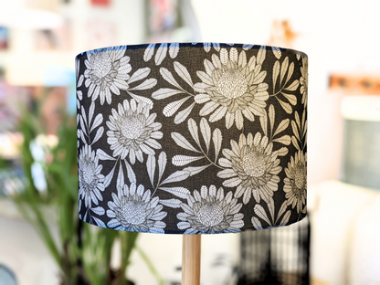 A close-up of the Emotive Collection’s Australian Waratah Lampshade, handmade with a bold white floral pattern on black, large flowers and leaves in Jocelyn Prousts style. The lamp is indoors, background softened with plants and furniture.