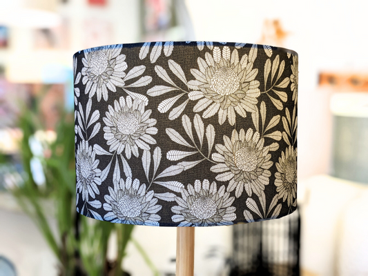 A close-up of the Emotive Collection’s Australian Waratah Lampshade, handmade with a bold white floral pattern on black, large flowers and leaves in Jocelyn Prousts style. The lamp is indoors, background softened with plants and furniture.