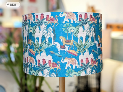 The Art Deco Jaipur Lampshade (35cm x 24cm) features vibrant elephants, horses, palm trees, and ornate buildings on a blue background, adding an exotic, royal, and tropical flair to your home.