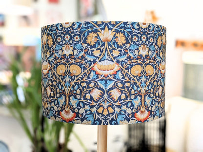 The Emotive Collection William Morris Lodden Lampshade is a cylindrical, handcrafted piece featuring a blue, gold, red, and cream floral pattern with a softly blurred background that highlights its artistic elegance.