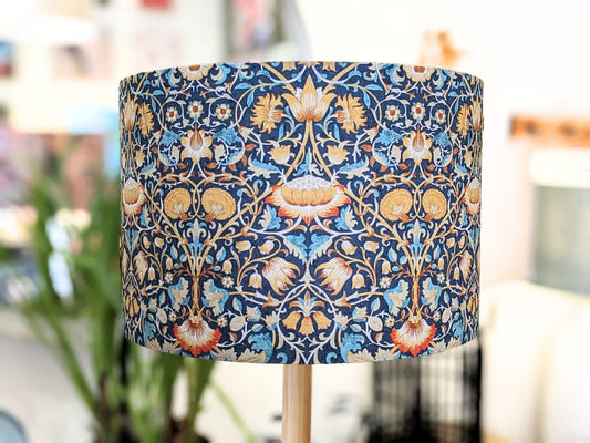 The Emotive Collection William Morris Lodden Lampshade is a cylindrical, handcrafted piece featuring a blue, gold, red, and cream floral pattern with a softly blurred background that highlights its artistic elegance.