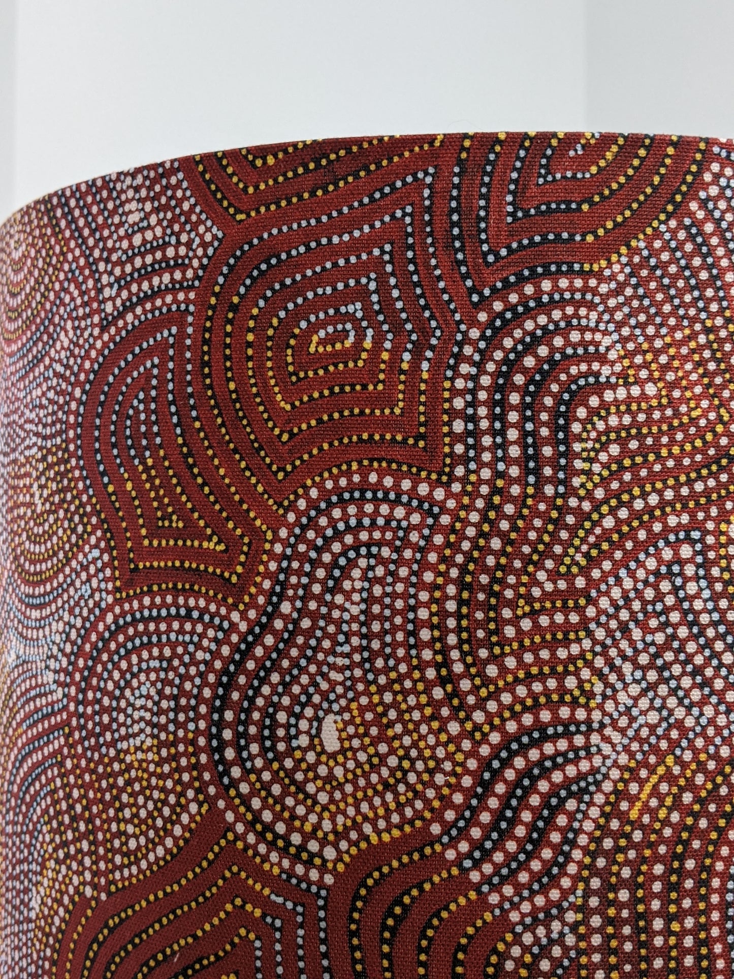 A close-up of the Warlu Mina Mina Jukurrpa (Ngalyipi) Indigenous Aboriginal lampshade by artist Mekeisha Napanangka Martin features red, white, yellow, and black curved lines and dots in a traditional dot painting style on a cylindrical surface.