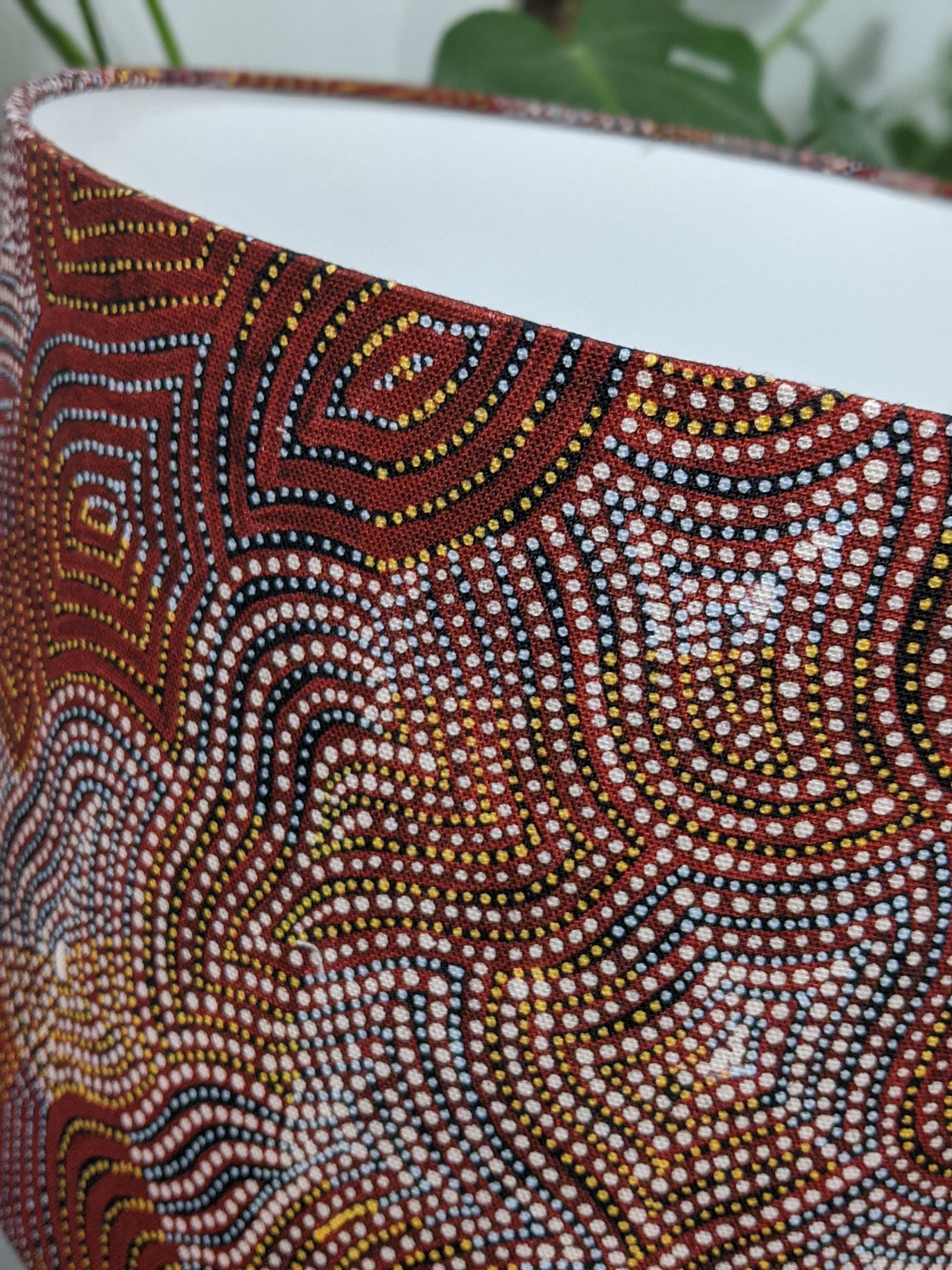 Close-up of the Warlu Mina Mina Jukurrpa (Ngalyipi) lampshade by Mekeisha Napanangka Martin, featuring intricate Aboriginal dot painting in red, yellow, black, and white with swirling patterns; green plant leaves blur in the background.