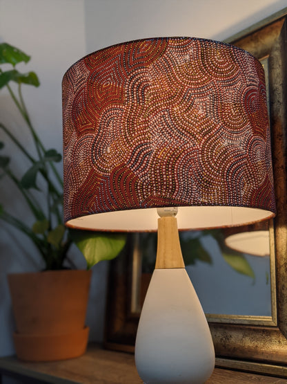 The Warlu Mina Mina Jukurrpa (Ngalyipi) table lamp by Mekeisha Napanangka Martin features an Indigenous Aboriginal design and a white base. It sits on a wooden surface with a leafy plant and framed mirror in the cozy background.