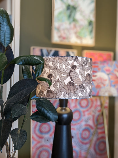 An Australian Cockatoo Lampshade by Emotive Collection, featuring handmade bird and leaf designs, accents a black lamp next to a leafy plant, with colorful abstract prints on a green wall in the background.
