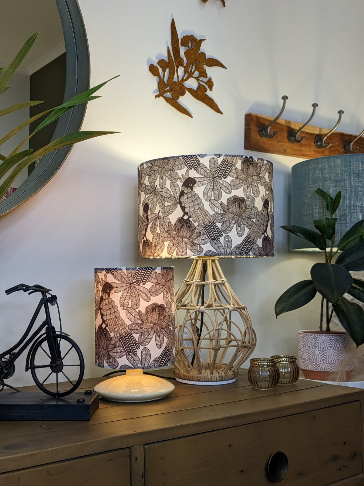 A cozy wooden sideboard displays a decorative bicycle, candle, two Emotive Collection Australian Cockatoo Lampshade lamps, potted plants, and gold candle holders. A mirror, wall hooks, and metal bird wall art hang above.