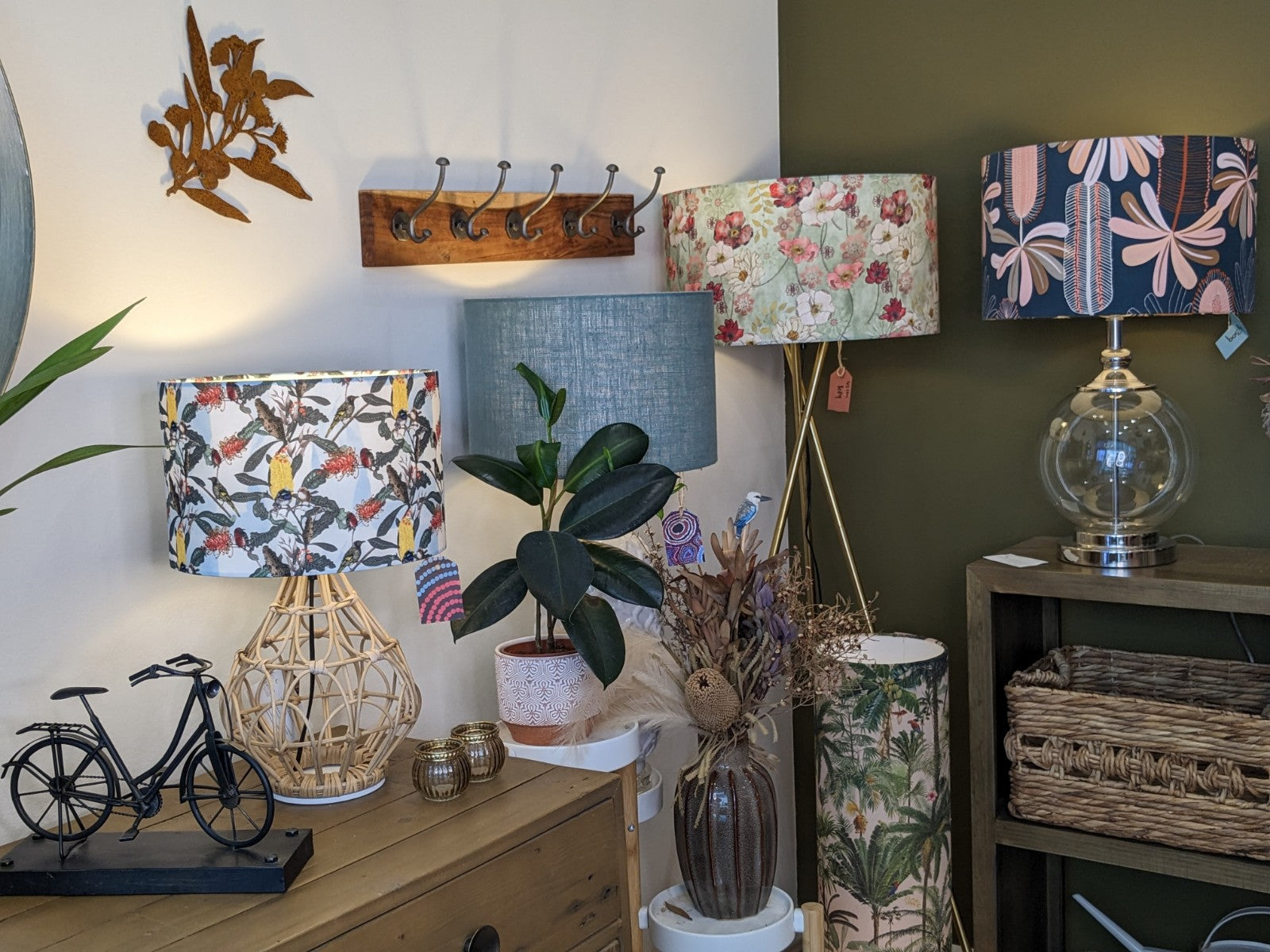 A cozy interior by Tamara Schneider features the Yellow Banksia & Native Birds Lampshade from the Emotive Collection, a potted plant, dried flowers, wicker baskets, a miniature bicycle, and a wall hook rack set against green and white walls.